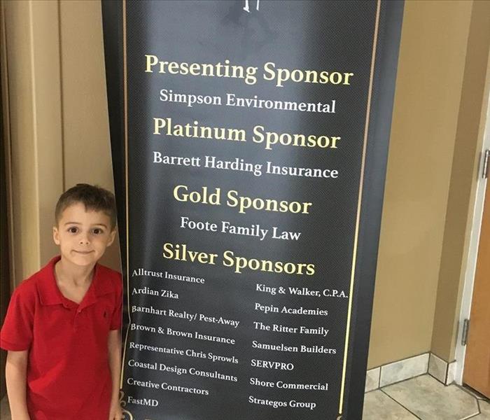 Brunette boy in red shirt standing by a black banner listing sponsors